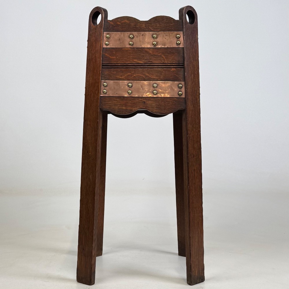An antique Arts and Crafts dark oak plant stand featuring copper straps with brass rivets, pierced handles, and scalloped edges.