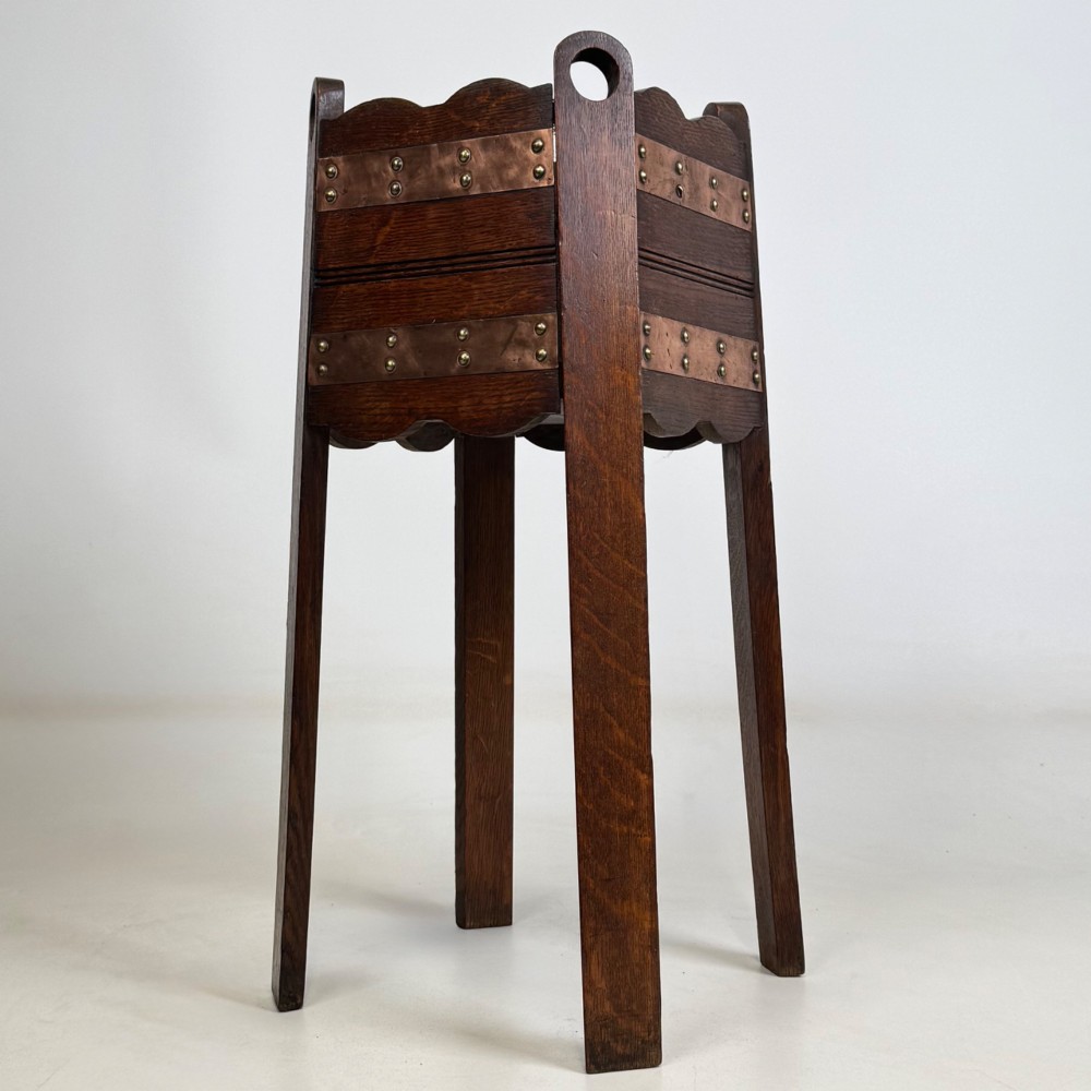An antique Arts and Crafts dark oak plant stand featuring copper straps with brass rivets, pierced handles, and scalloped edges. View IV