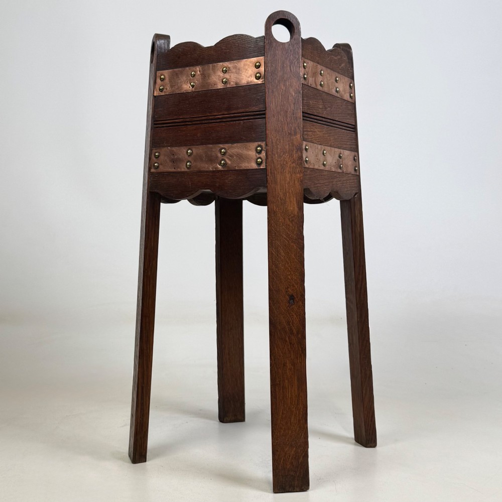 An antique Arts and Crafts dark oak plant stand featuring copper straps with brass rivets, pierced handles, and scalloped edges. View VI