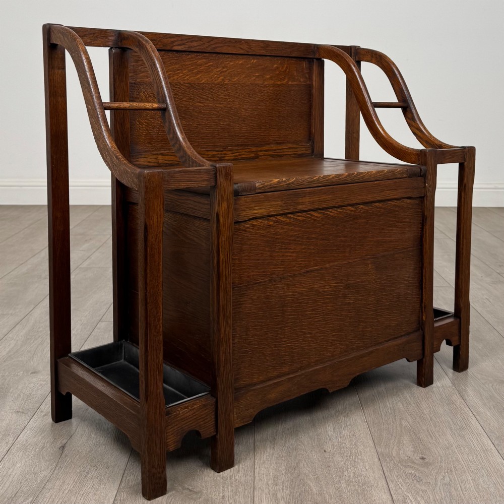 An antique Edwardian golden oak hall seat featuring a central panelled backrest and integrated side umbrella stands with metal drip trays. View VIII