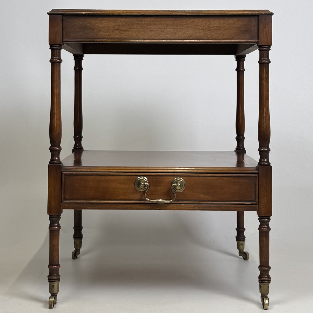 A pair of 20th-century warm mahogany bedside tables featuring turned support pillars, open middle shelves, single lower drawers with brass handles, and brass castors. View II