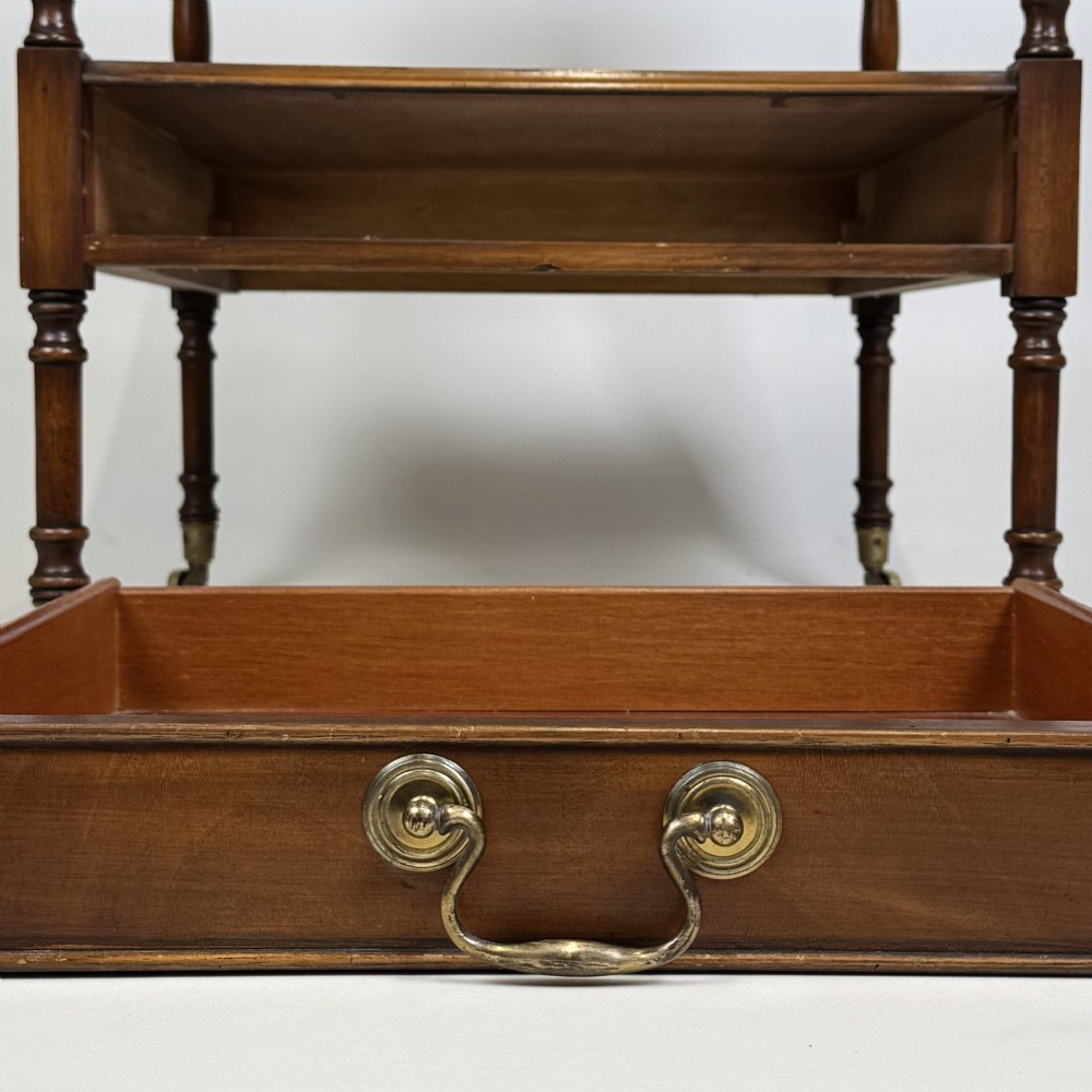 A pair of 20th-century warm mahogany bedside tables featuring turned support pillars, open middle shelves, single lower drawers with brass handles, and brass castors. View XI