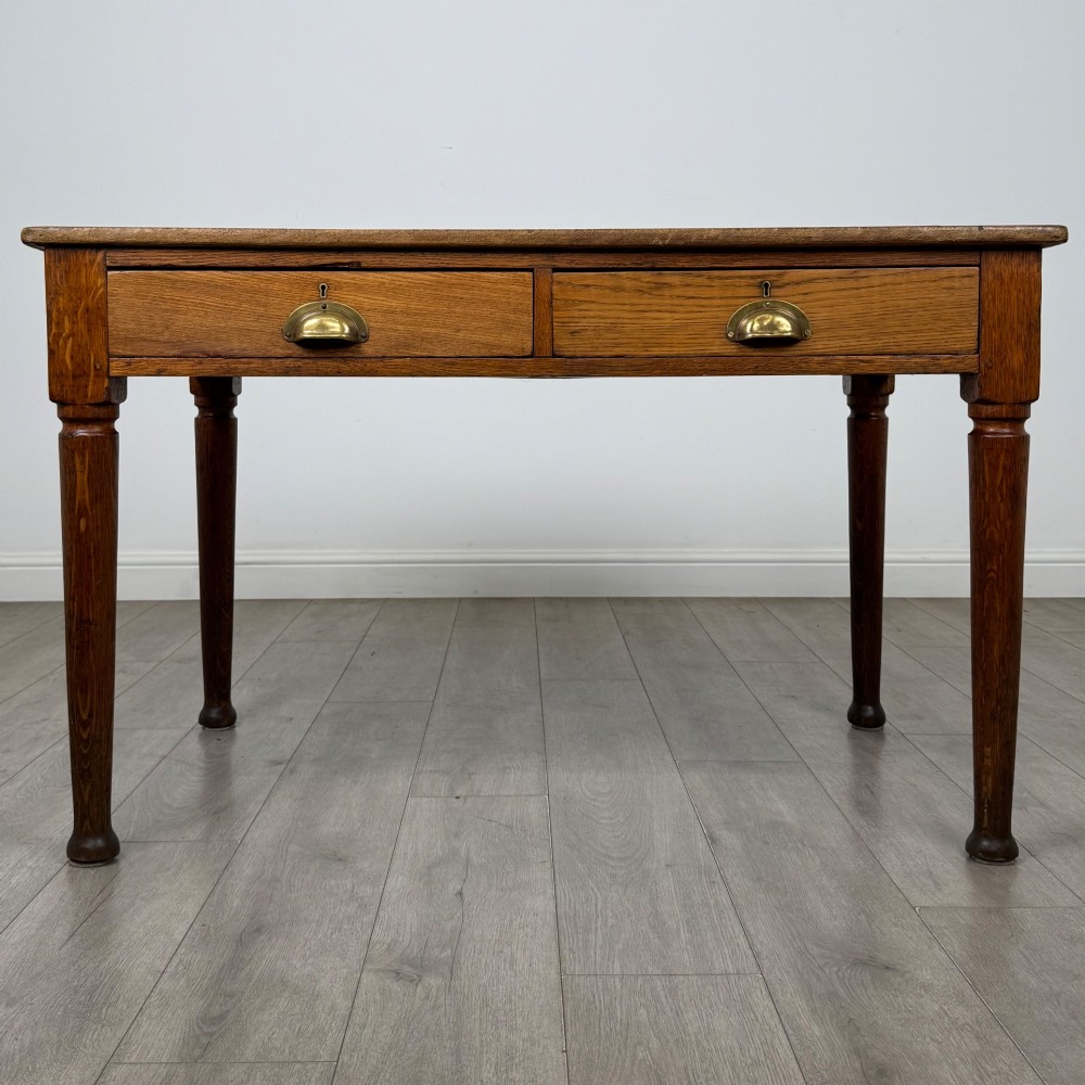 An antique early 20th-century oak writing table featuring two front drawers with brass cup handles and turned legs.