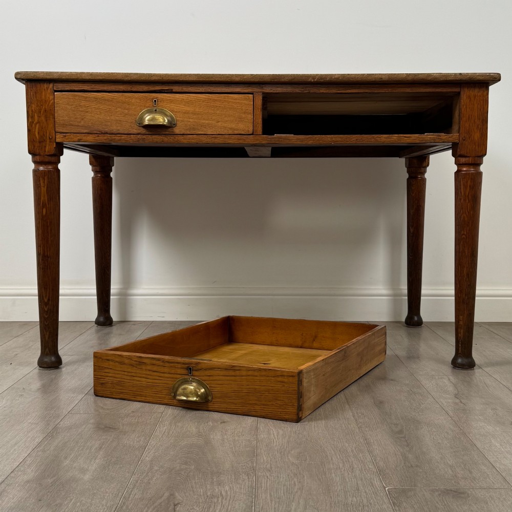 An antique early 20th-century oak writing table featuring two front drawers with brass cup handles and turned legs. View X