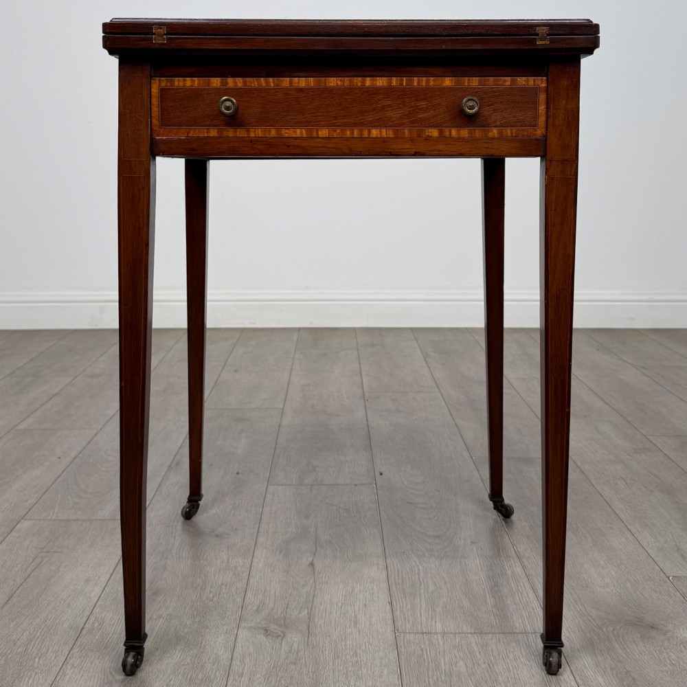 An Edwardian mahogany envelope card table with crossbanded satinwood inlay, a single drawer, and tapered square legs on brass castors.