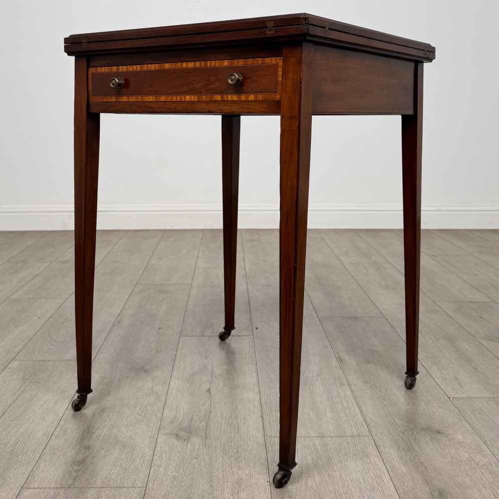 An Edwardian mahogany envelope card table with crossbanded satinwood inlay, a single drawer, and tapered square legs on brass castors. View II