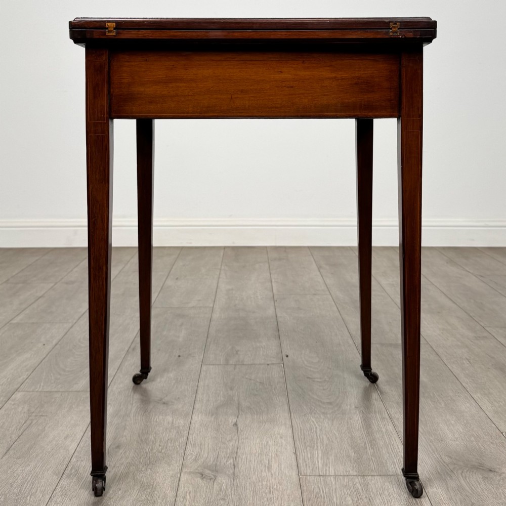 An Edwardian mahogany envelope card table with crossbanded satinwood inlay, a single drawer, and tapered square legs on brass castors. View III