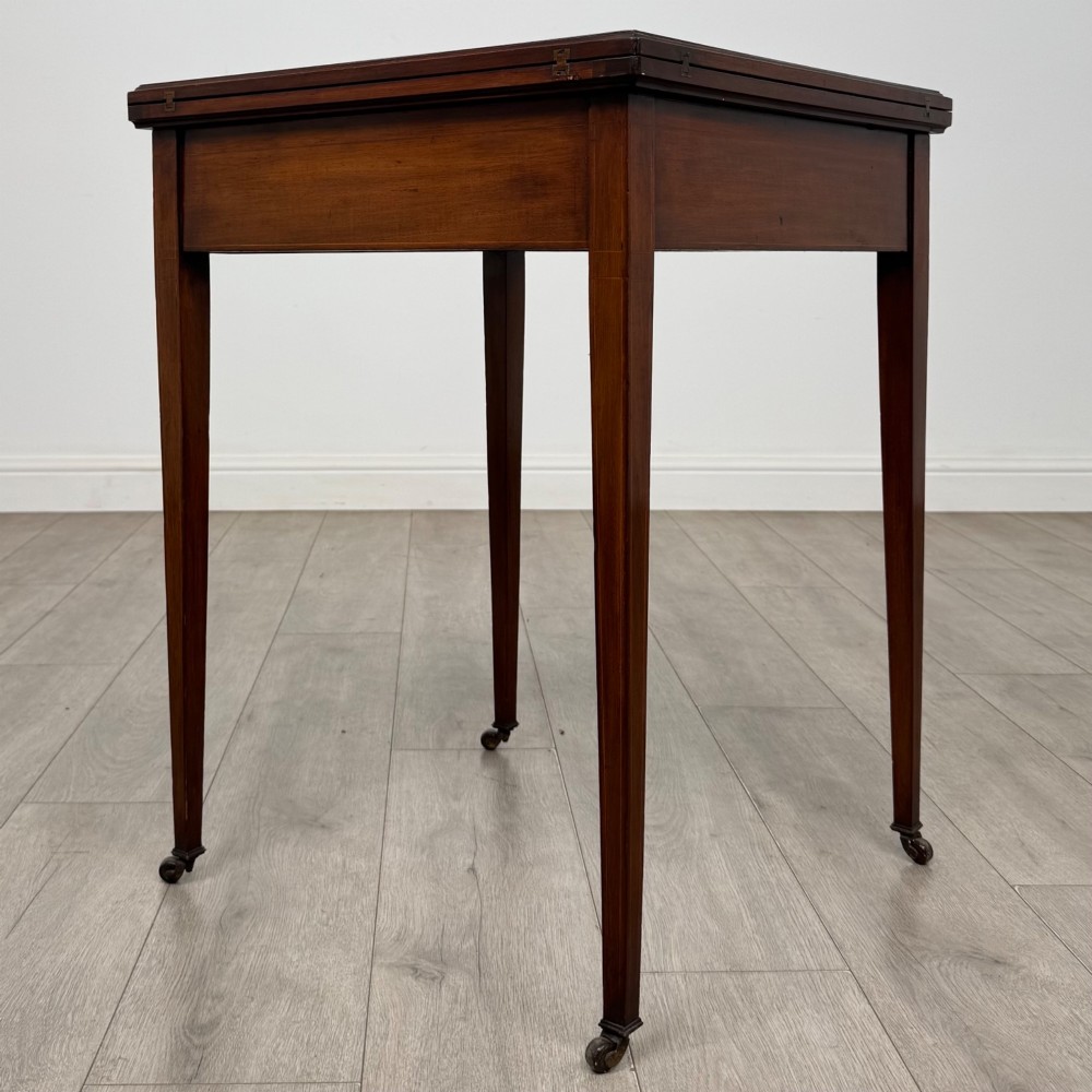 An Edwardian mahogany envelope card table with crossbanded satinwood inlay, a single drawer, and tapered square legs on brass castors. View IV