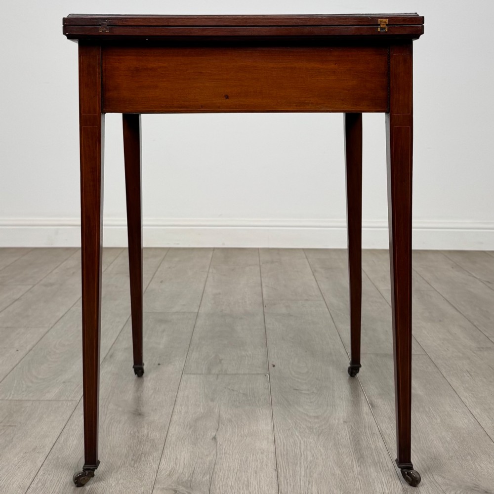 An Edwardian mahogany envelope card table with crossbanded satinwood inlay, a single drawer, and tapered square legs on brass castors. View V