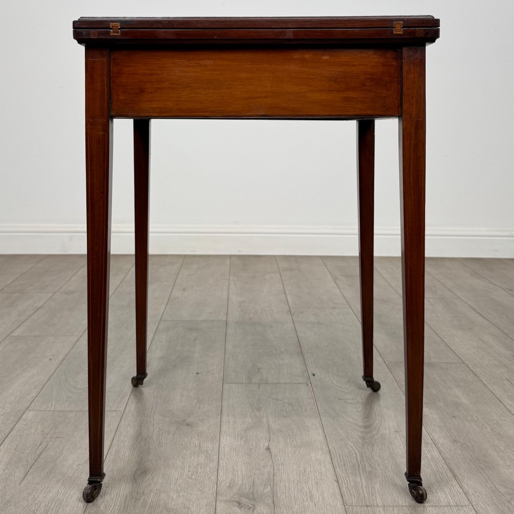 An Edwardian mahogany envelope card table with crossbanded satinwood inlay, a single drawer, and tapered square legs on brass castors. View VII