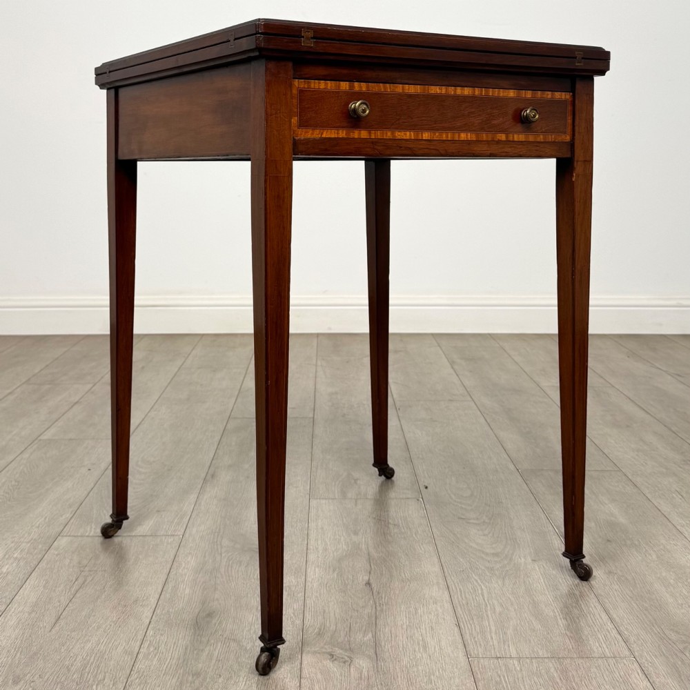 An Edwardian mahogany envelope card table with crossbanded satinwood inlay, a single drawer, and tapered square legs on brass castors. View VIII