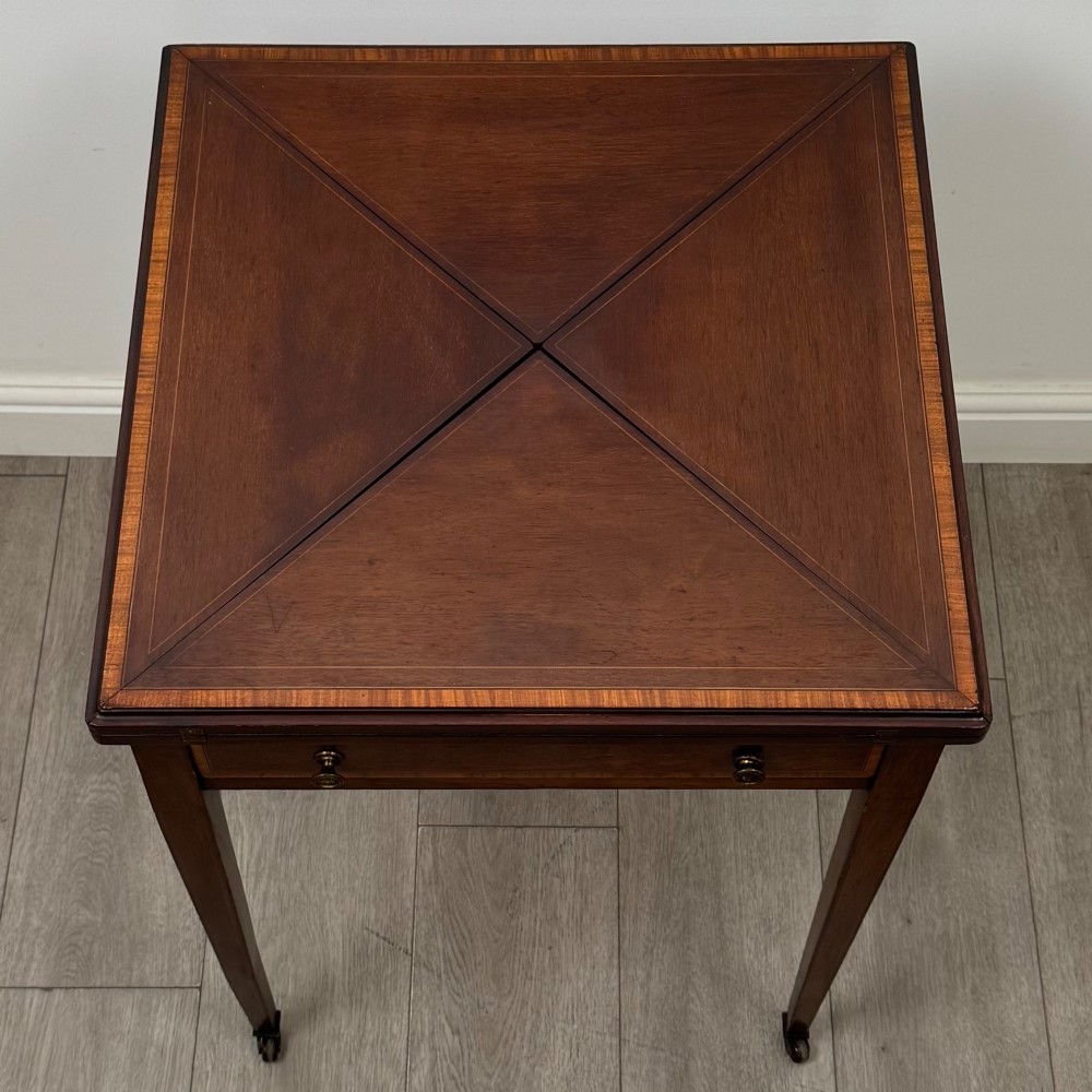 An Edwardian mahogany envelope card table with crossbanded satinwood inlay, a single drawer, and tapered square legs on brass castors. View IX