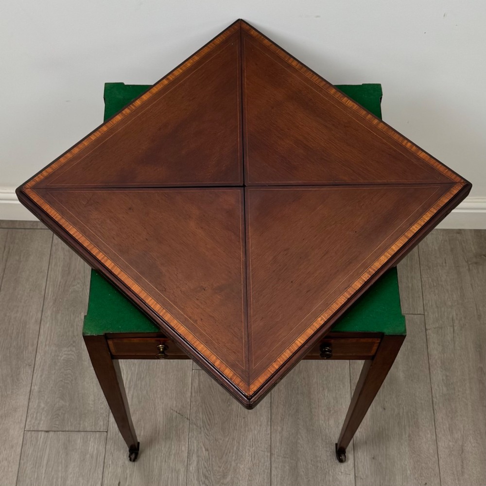 An Edwardian mahogany envelope card table with crossbanded satinwood inlay, a single drawer, and tapered square legs on brass castors. View X