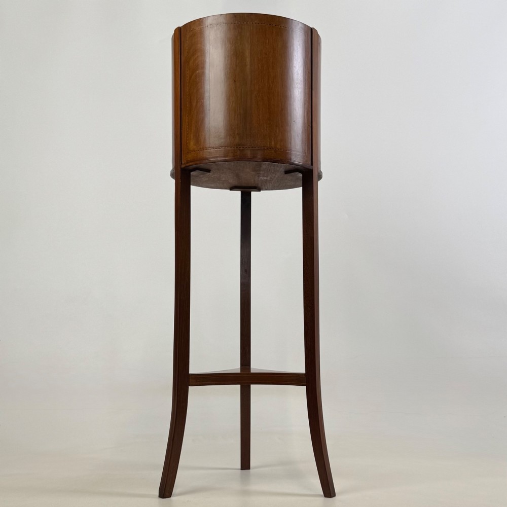 A tall Edwardian mahogany plant stand featuring a cylindrical top bucket with subtle line inlay, supported by three elongated curved legs and a triangular lower shelf.