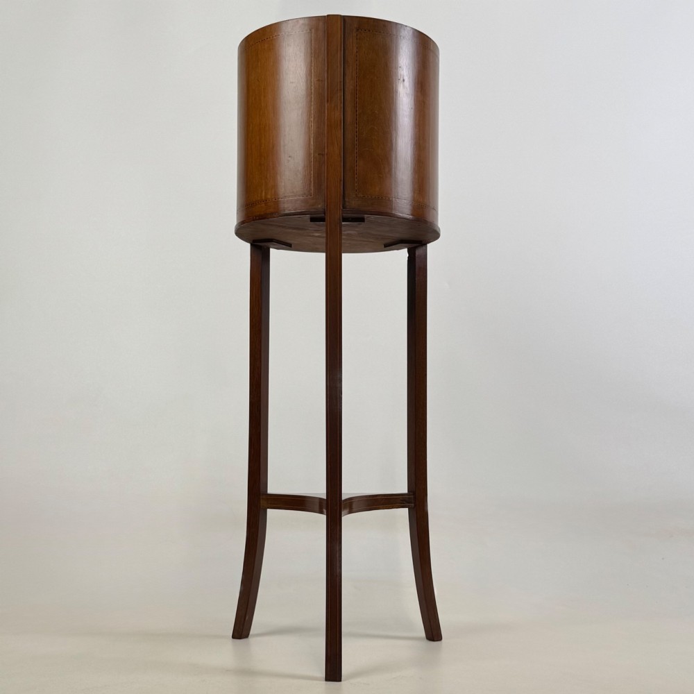 A tall Edwardian mahogany plant stand featuring a cylindrical top bucket with subtle line inlay, supported by three elongated curved legs and a triangular lower shelf. View III