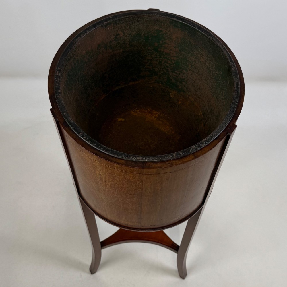 A tall Edwardian mahogany plant stand featuring a cylindrical top bucket with subtle line inlay, supported by three elongated curved legs and a triangular lower shelf. View V