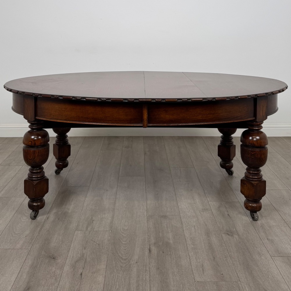 A large antique Edwardian extending oak dining table with detailed carved borders, thick turned legs, and brass castors. View VI