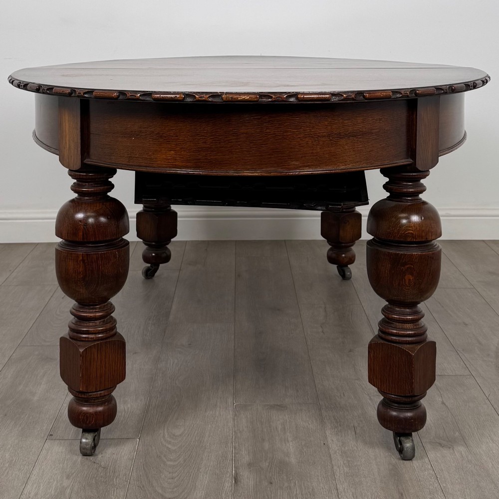 A large antique Edwardian extending oak dining table with detailed carved borders, thick turned legs, and brass castors. View VII