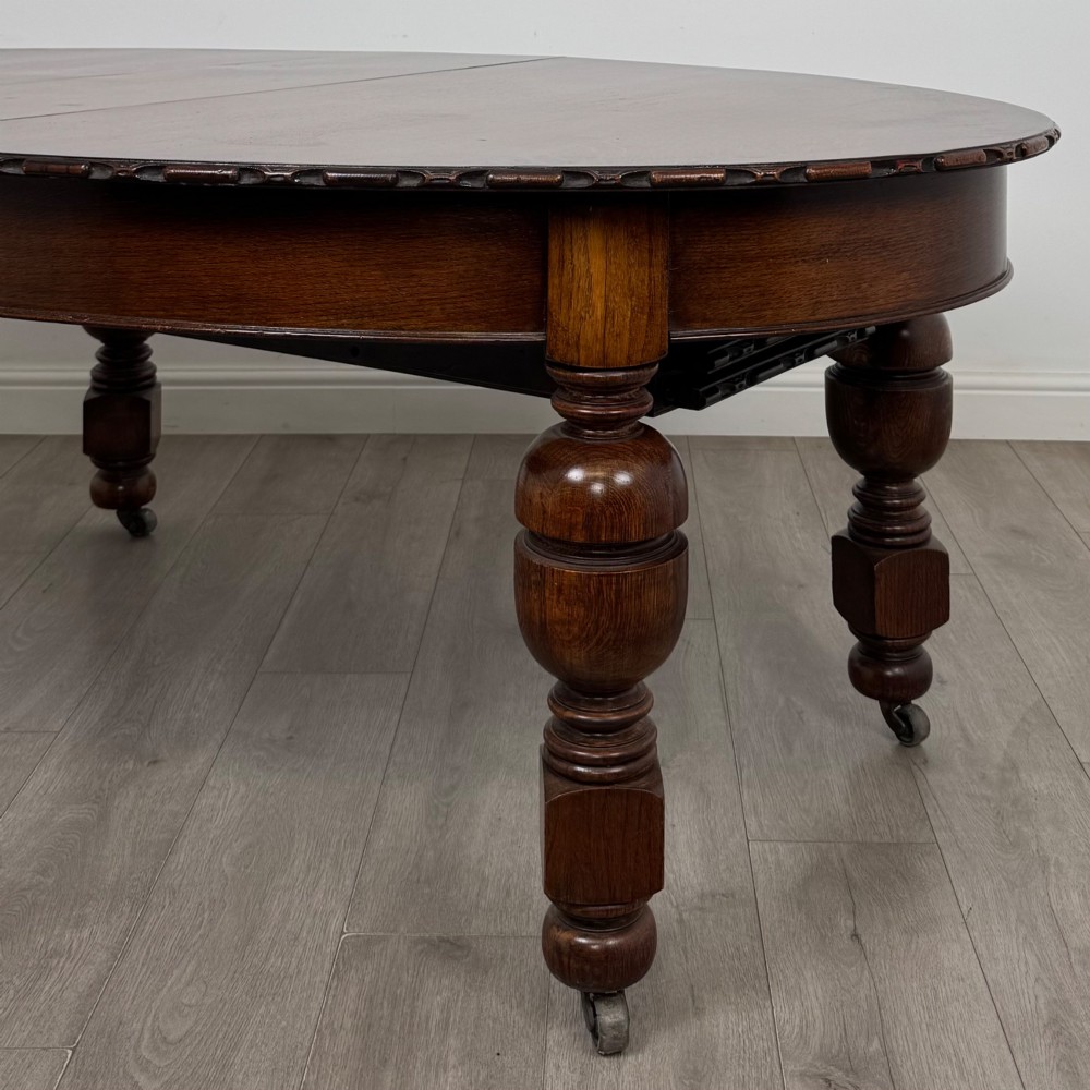 A large antique Edwardian extending oak dining table with detailed carved borders, thick turned legs, and brass castors. View IX