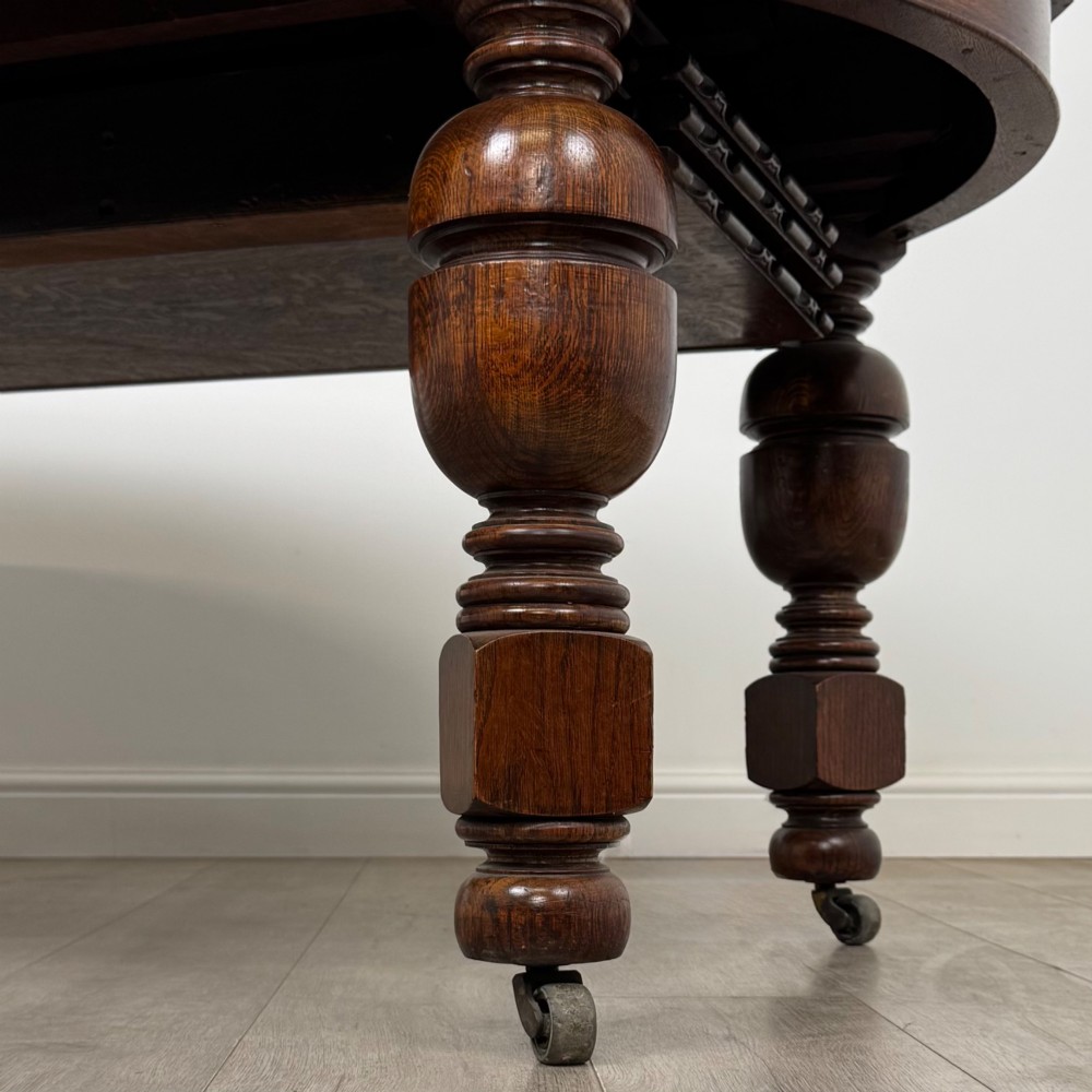 A large antique Edwardian extending oak dining table with detailed carved borders, thick turned legs, and brass castors. View X