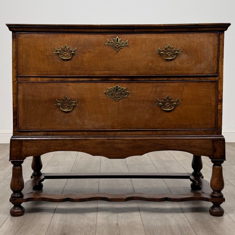 An antique 19th-century William and Mary style walnut chest of drawers featuring two deep drawers with ornate brass drop handles, raised on turned legs connected by a wavy stretcher base.