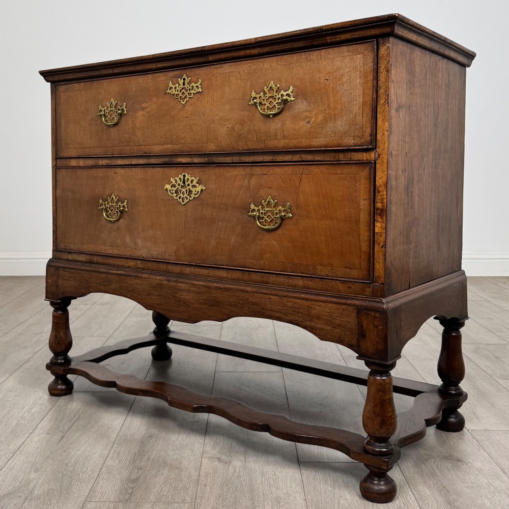 An antique 19th-century William and Mary style walnut chest of drawers featuring two deep drawers with ornate brass drop handles, raised on turned legs connected by a wavy stretcher base. View II