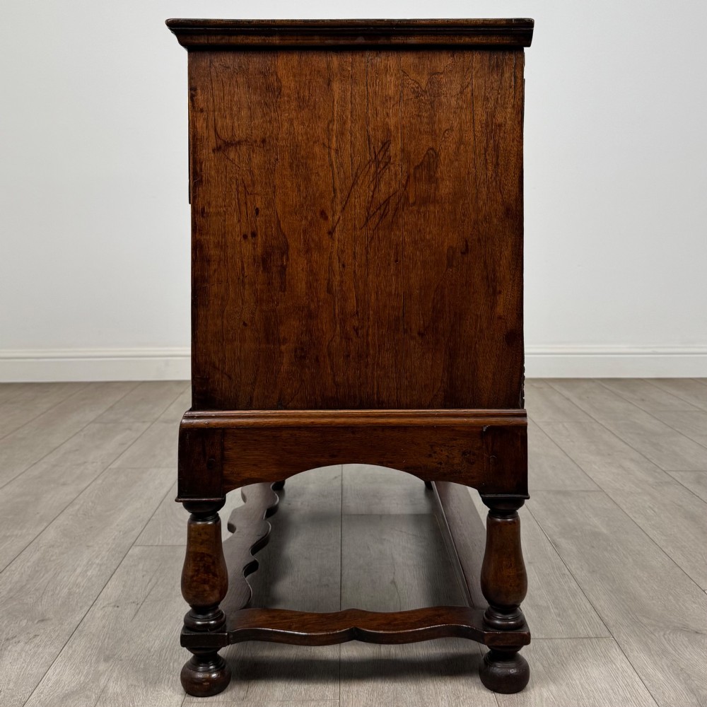 An antique 19th-century William and Mary style walnut chest of drawers featuring two deep drawers with ornate brass drop handles, raised on turned legs connected by a wavy stretcher base. View III