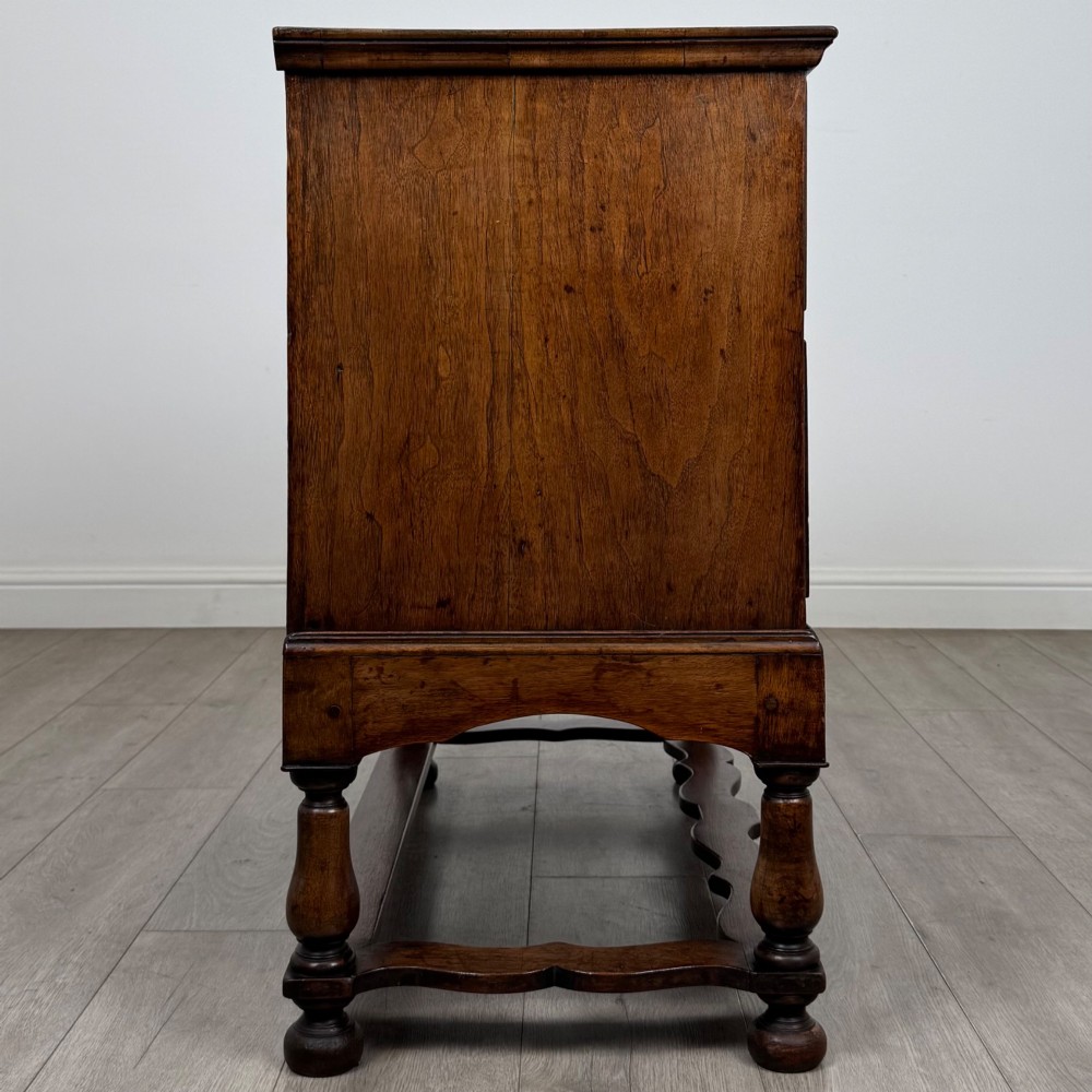 An antique 19th-century William and Mary style walnut chest of drawers featuring two deep drawers with ornate brass drop handles, raised on turned legs connected by a wavy stretcher base. View VII