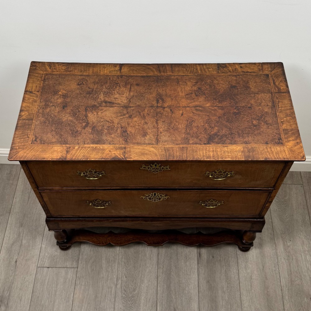 An antique 19th-century William and Mary style walnut chest of drawers featuring two deep drawers with ornate brass drop handles, raised on turned legs connected by a wavy stretcher base. View IX