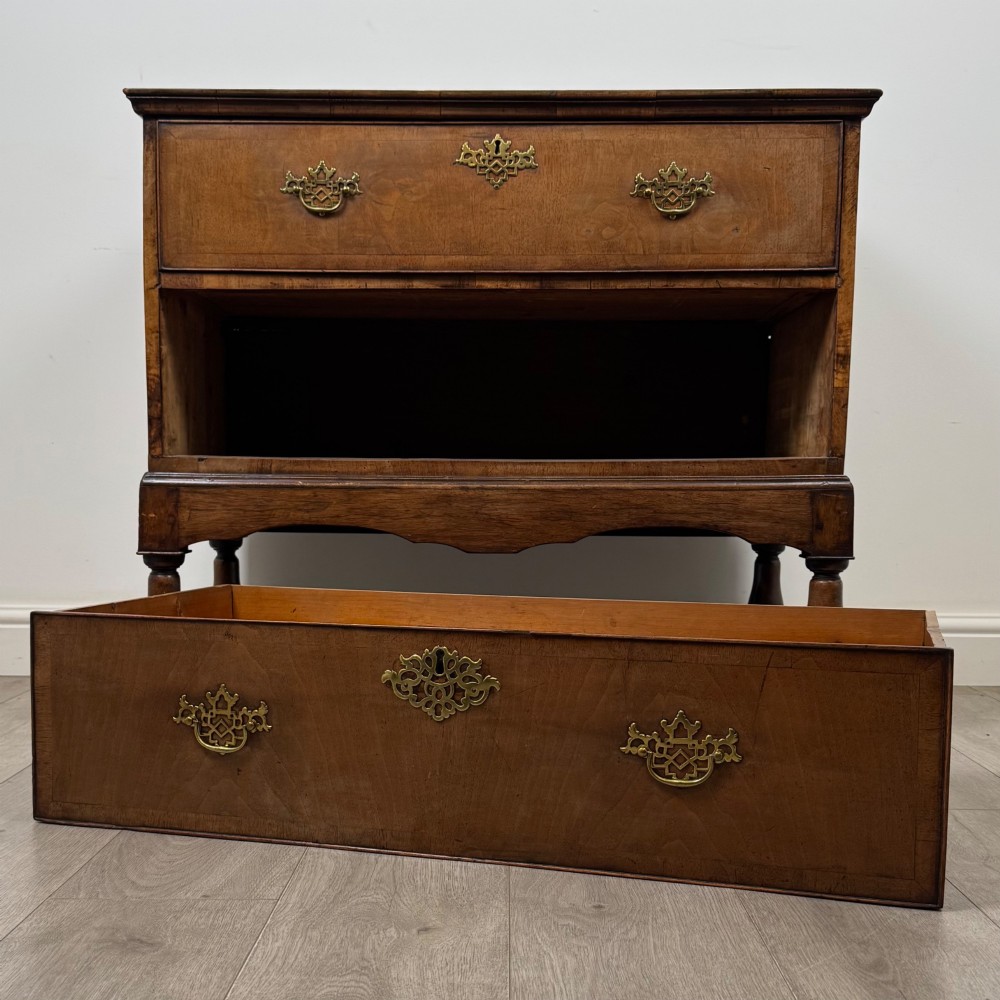 An antique 19th-century William and Mary style walnut chest of drawers featuring two deep drawers with ornate brass drop handles, raised on turned legs connected by a wavy stretcher base. View X