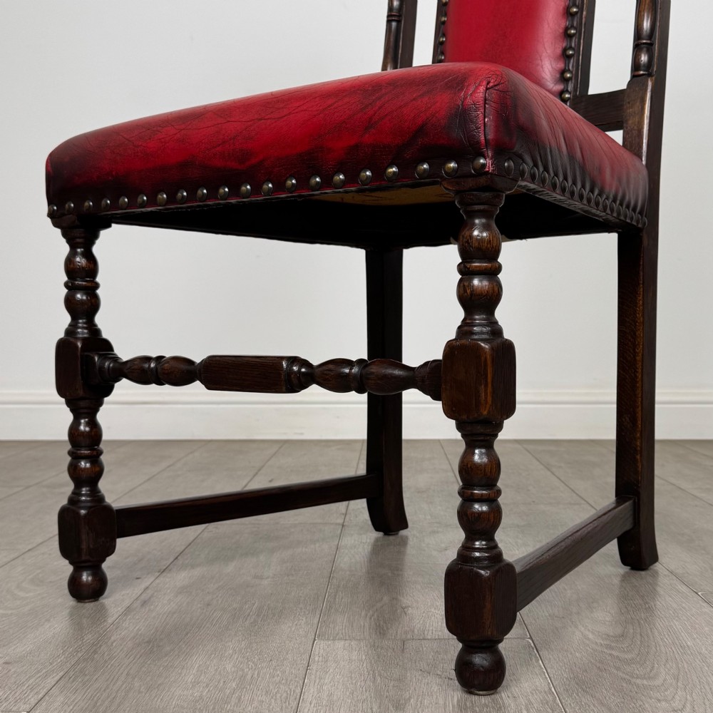 A set of six dark oak dining chairs featuring red leather upholstery, carved crests, turned legs, and brass stud trimming. View VI