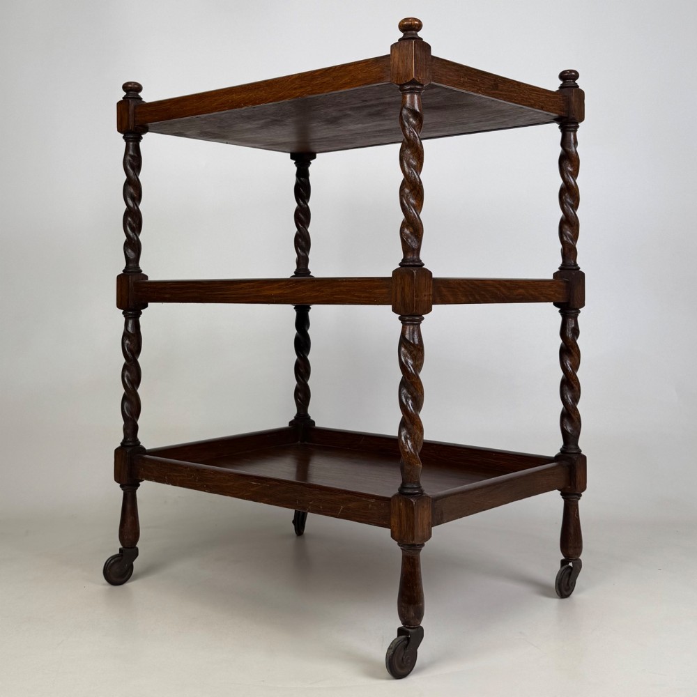 A three-tier dark oak tea trolley featuring spiralled barley twist corner supports, turned top finials, and castor wheels. View II