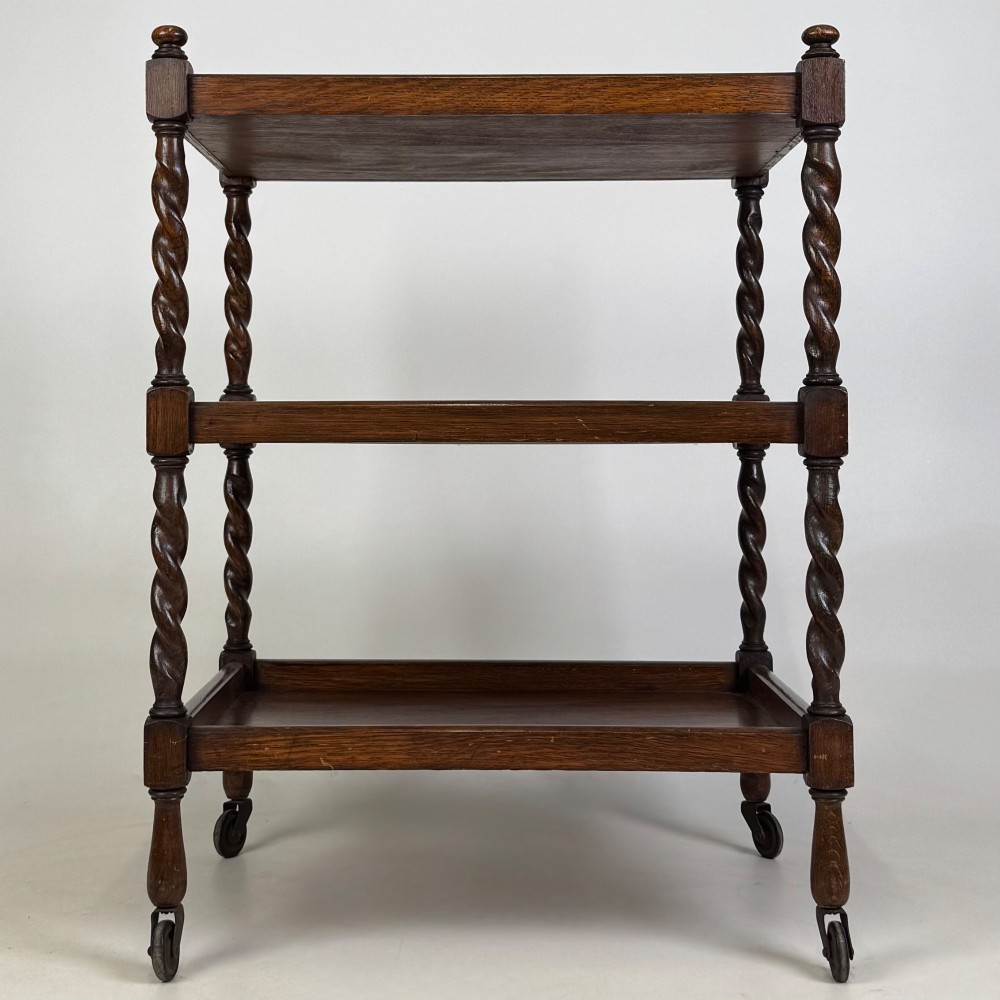 A three-tier dark oak tea trolley featuring spiralled barley twist corner supports, turned top finials, and castor wheels. View V