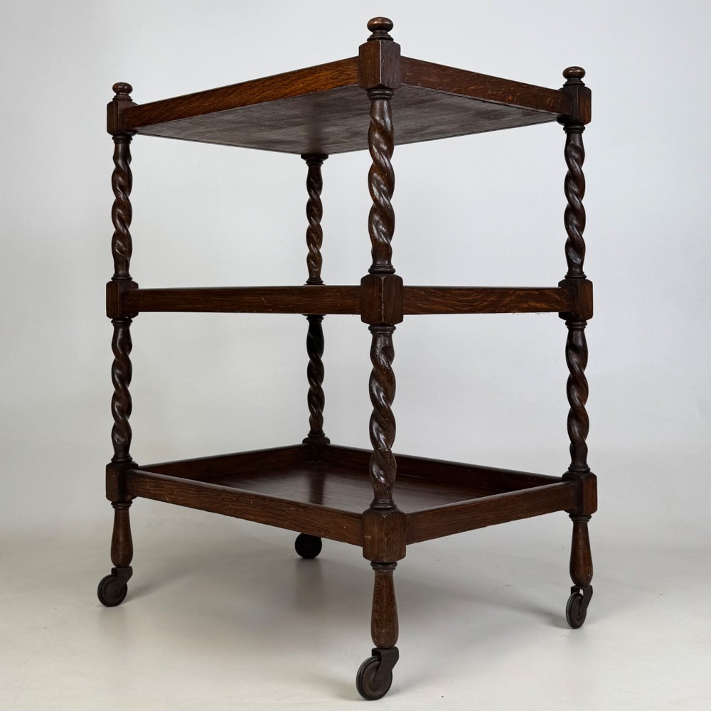 A three-tier dark oak tea trolley featuring spiralled barley twist corner supports, turned top finials, and castor wheels. View VI