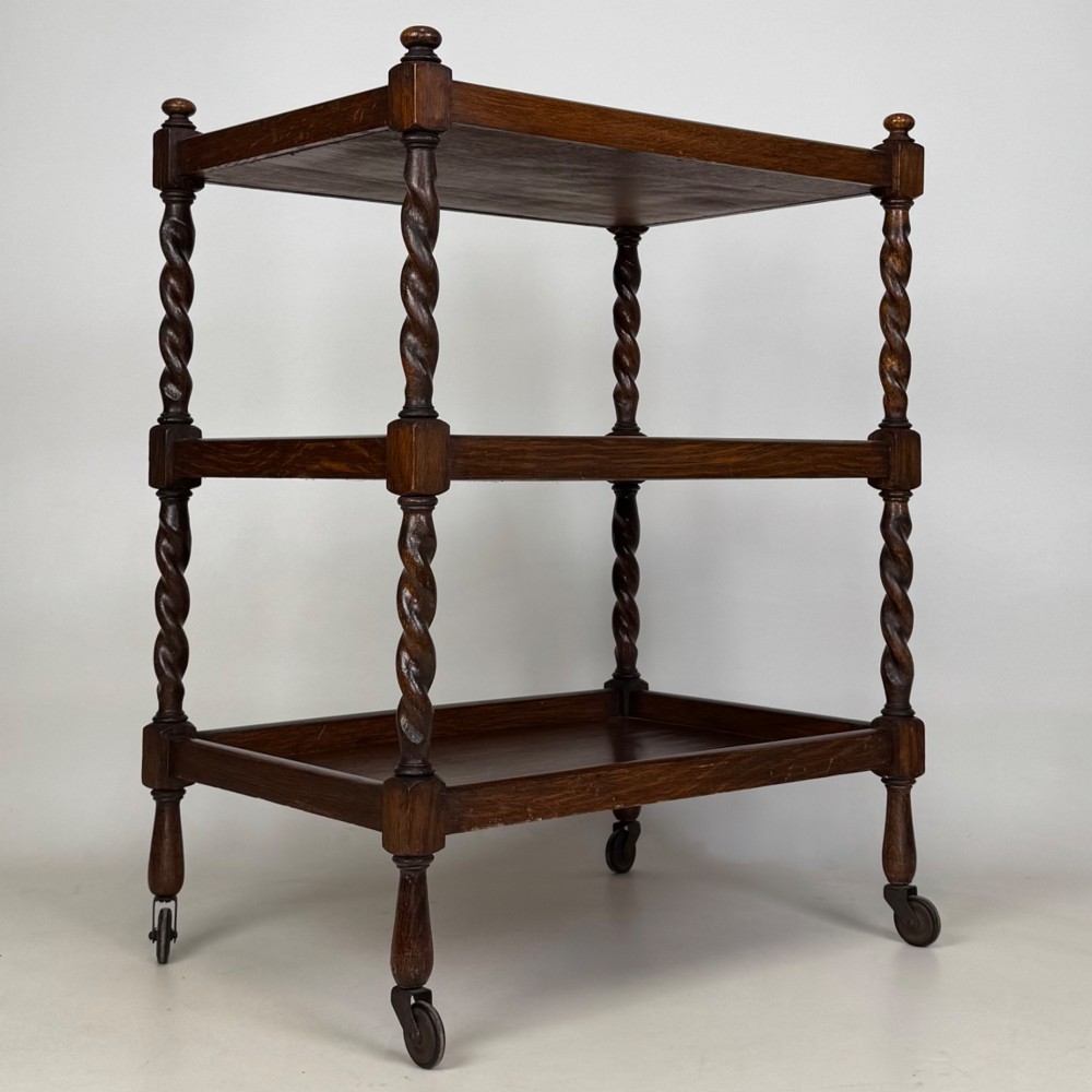 A three-tier dark oak tea trolley featuring spiralled barley twist corner supports, turned top finials, and castor wheels. View VIII