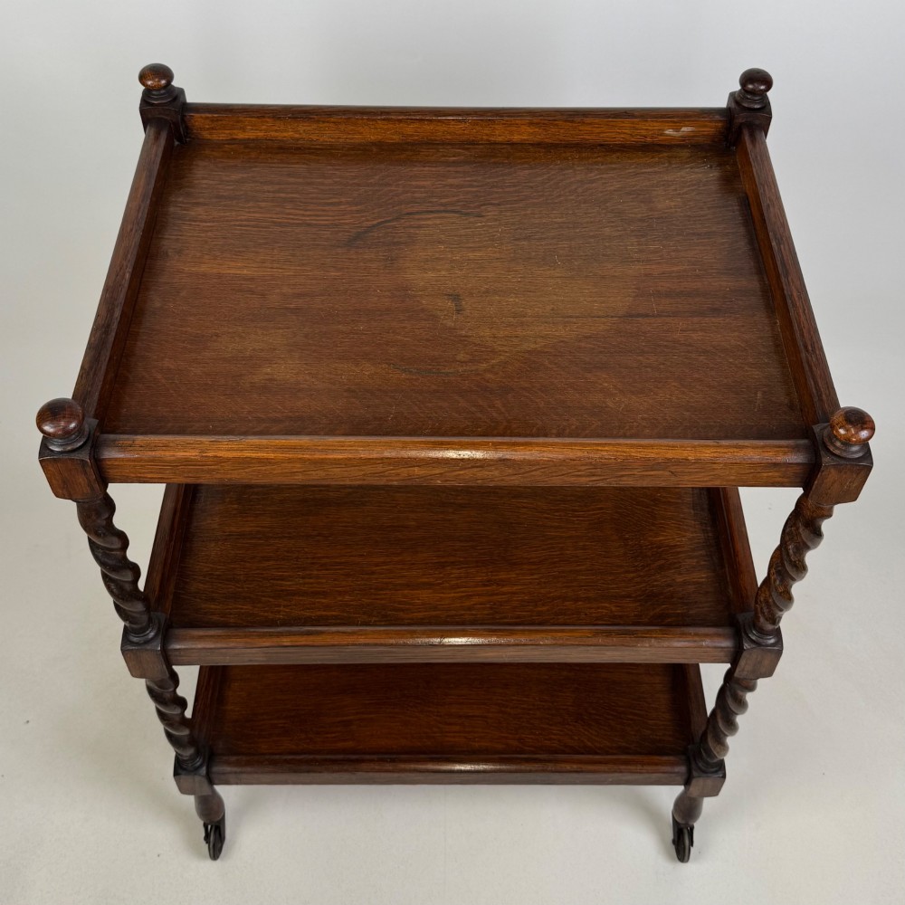 A three-tier dark oak tea trolley featuring spiralled barley twist corner supports, turned top finials, and castor wheels. View IX