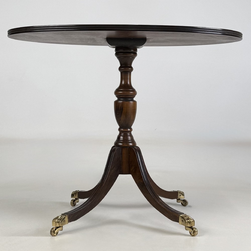 A dark brown oak pedestal side table with an oval top, turned central column, and swept legs featuring brass paw castors.