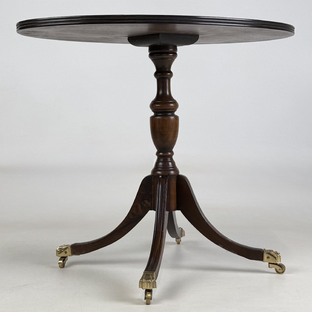A dark brown oak pedestal side table with an oval top, turned central column, and swept legs featuring brass paw castors. View IV