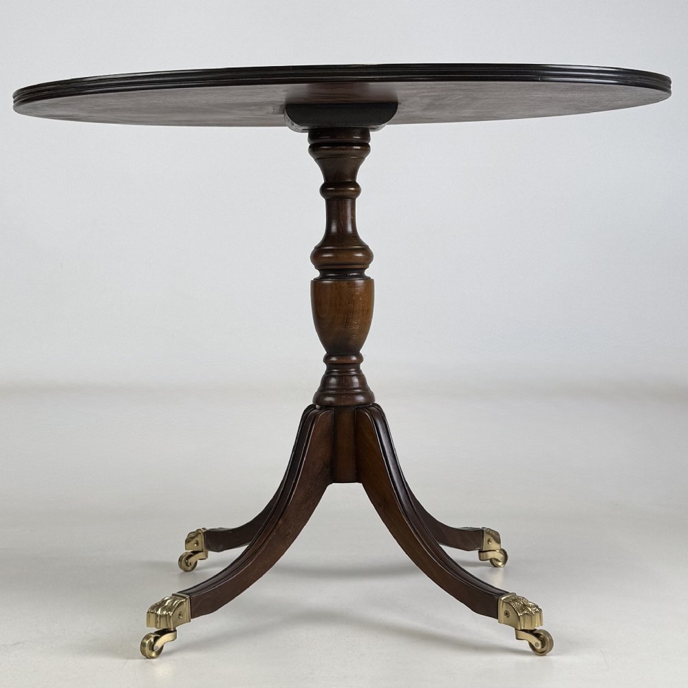 A dark brown oak pedestal side table with an oval top, turned central column, and swept legs featuring brass paw castors. View V