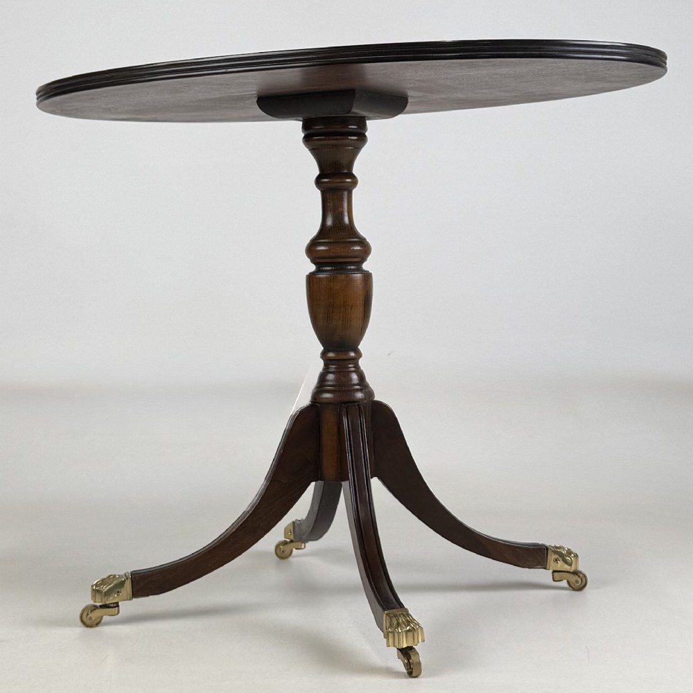 A dark brown oak pedestal side table with an oval top, turned central column, and swept legs featuring brass paw castors. View VI