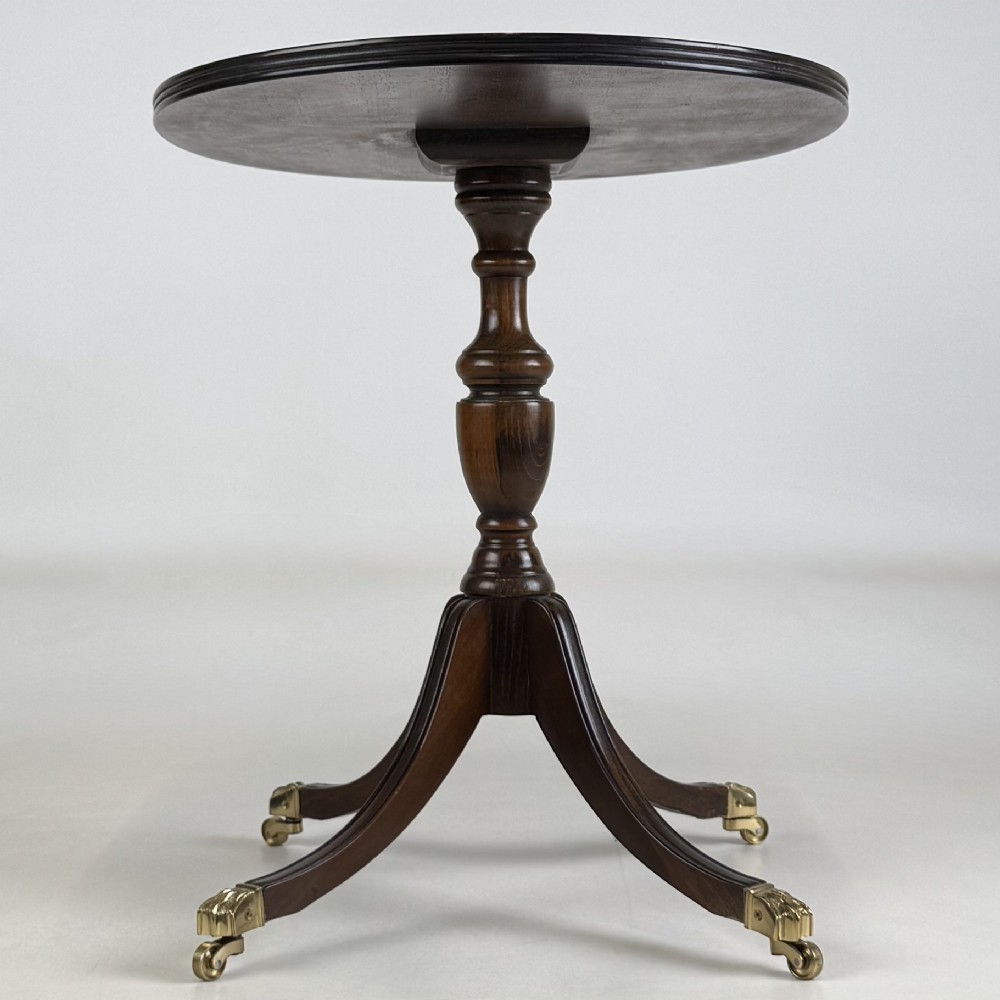 A dark brown oak pedestal side table with an oval top, turned central column, and swept legs featuring brass paw castors. View VII