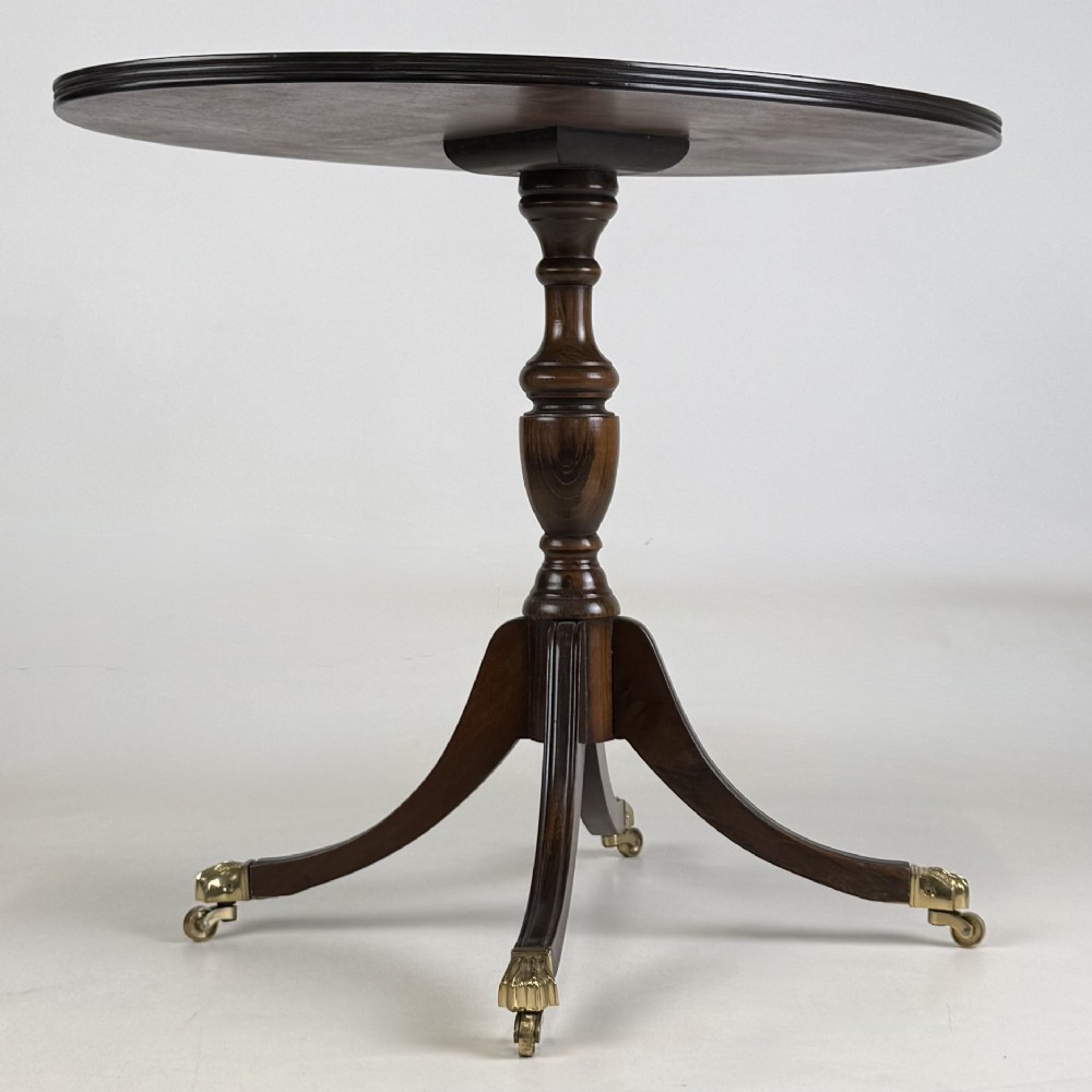 A dark brown oak pedestal side table with an oval top, turned central column, and swept legs featuring brass paw castors. View VIII