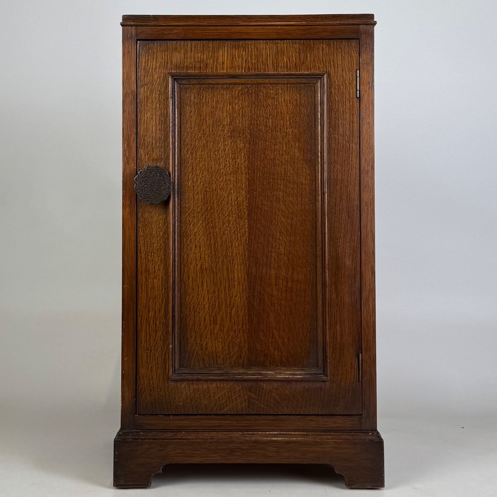 A standing antique dark oak bedside cabinet with a single framed door, a round carved handle, and a solid plinth base.
