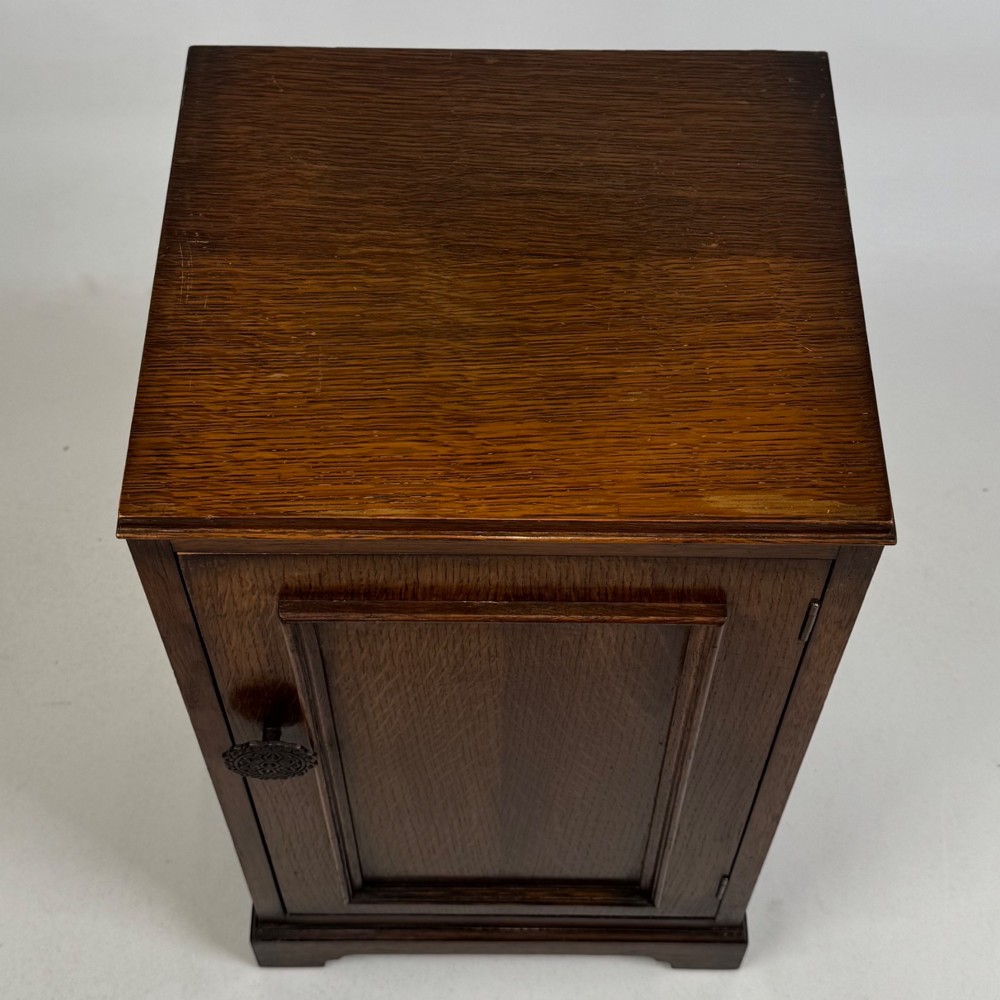 A standing antique dark oak bedside cabinet with a single framed door, a round carved handle, and a solid plinth base. View X