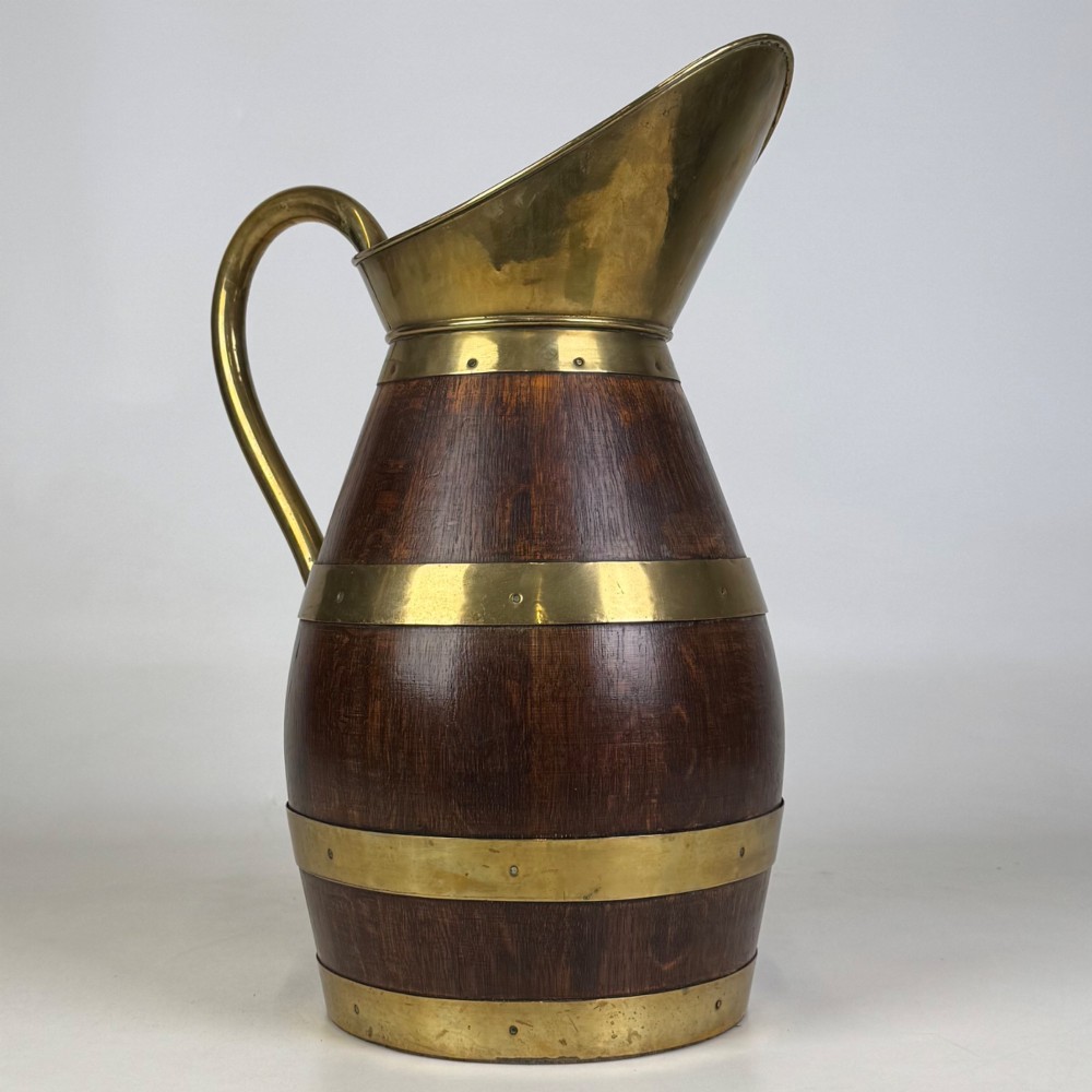 A tall dark oak wooden jug styled as an umbrella stand, featuring decorative brass bands, a curved brass handle, and a brass spout. View VII