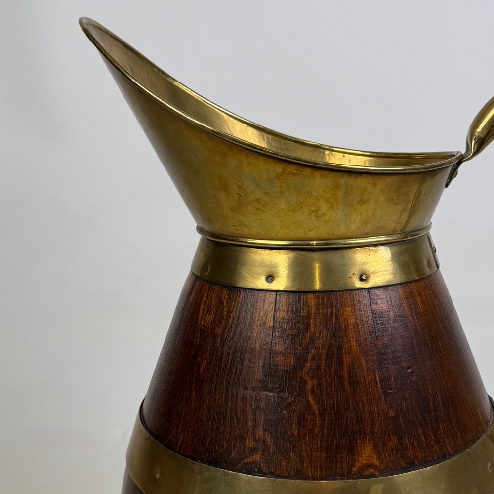 A tall dark oak wooden jug styled as an umbrella stand, featuring decorative brass bands, a curved brass handle, and a brass spout. View X