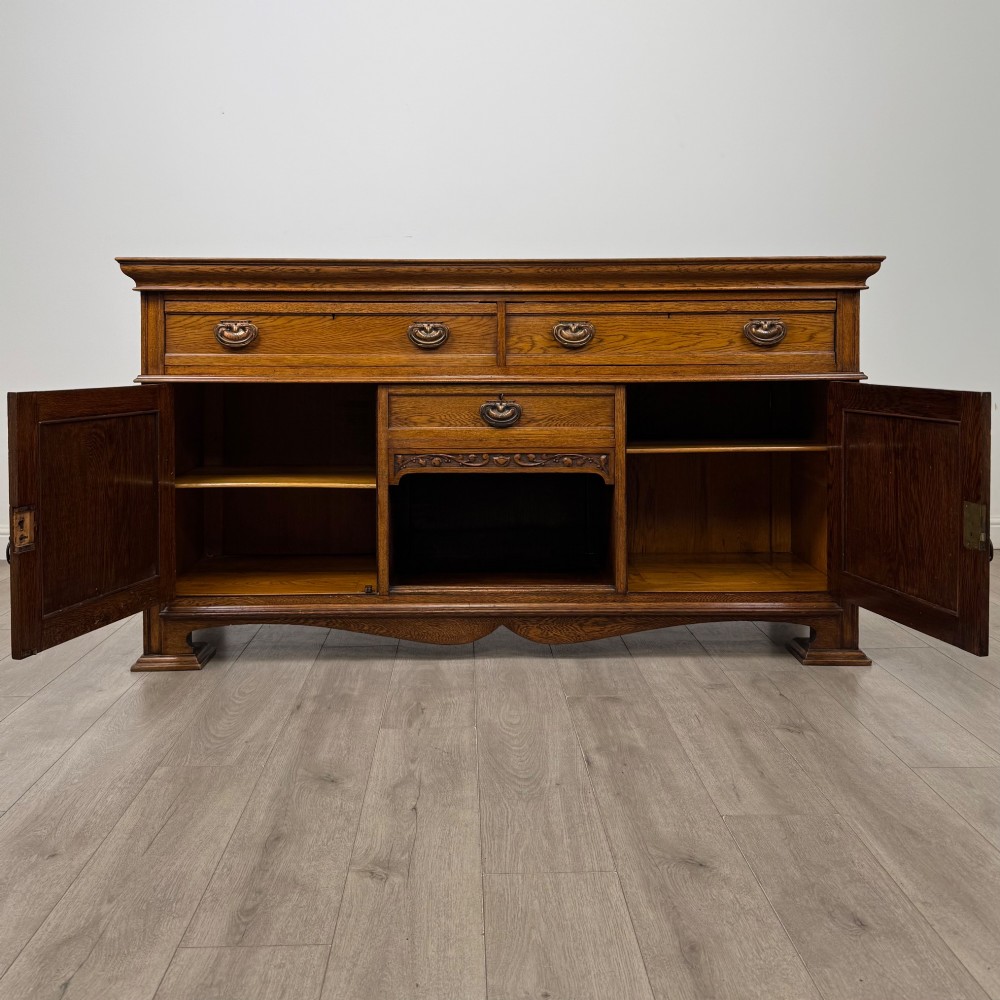 An early 20th-century oak Arts and Crafts sideboard featuring hand-carved foliate panels, copper drop handles, multiple drawers, and a central open recess. View II