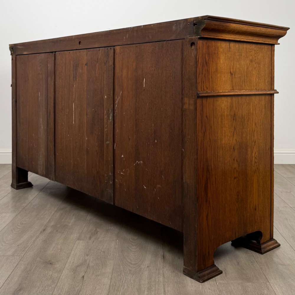 An early 20th-century oak Arts and Crafts sideboard featuring hand-carved foliate panels, copper drop handles, multiple drawers, and a central open recess. View VII