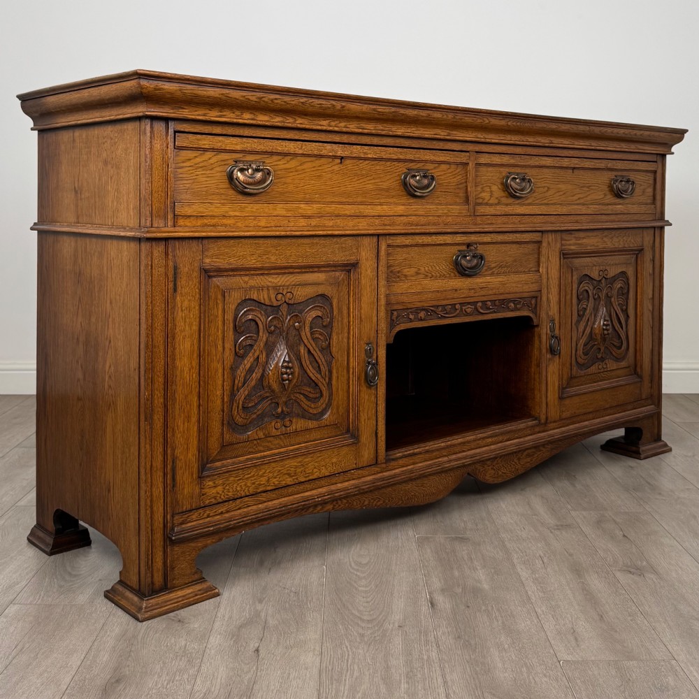 An early 20th-century oak Arts and Crafts sideboard featuring hand-carved foliate panels, copper drop handles, multiple drawers, and a central open recess. View IX