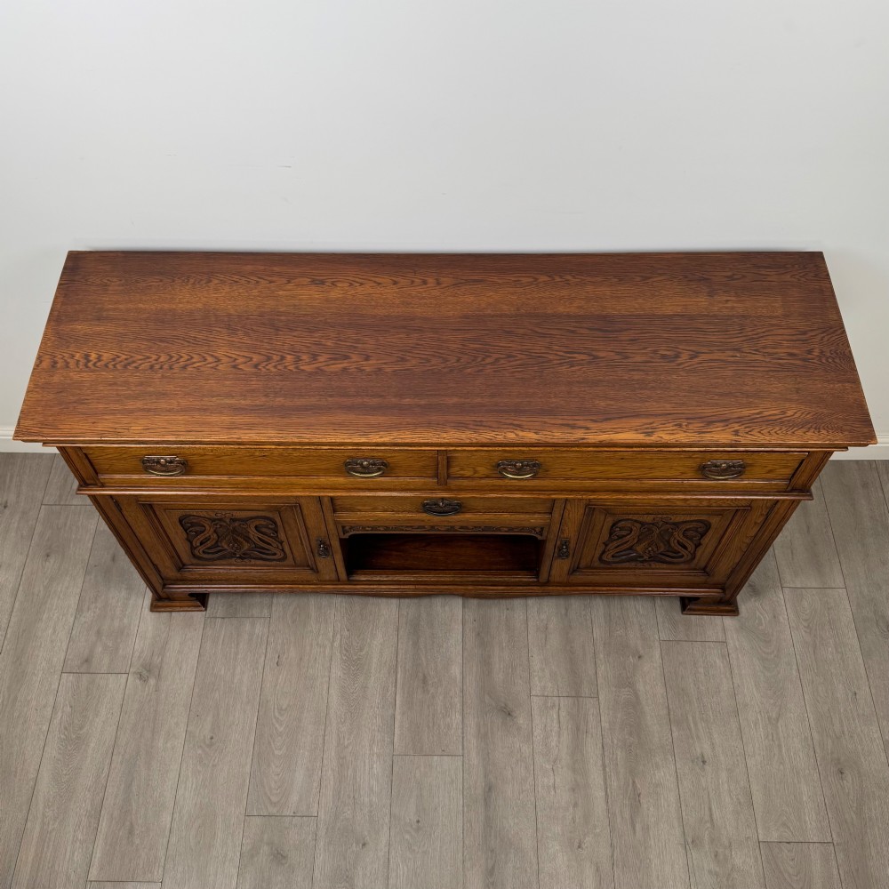 An early 20th-century oak Arts and Crafts sideboard featuring hand-carved foliate panels, copper drop handles, multiple drawers, and a central open recess. View X